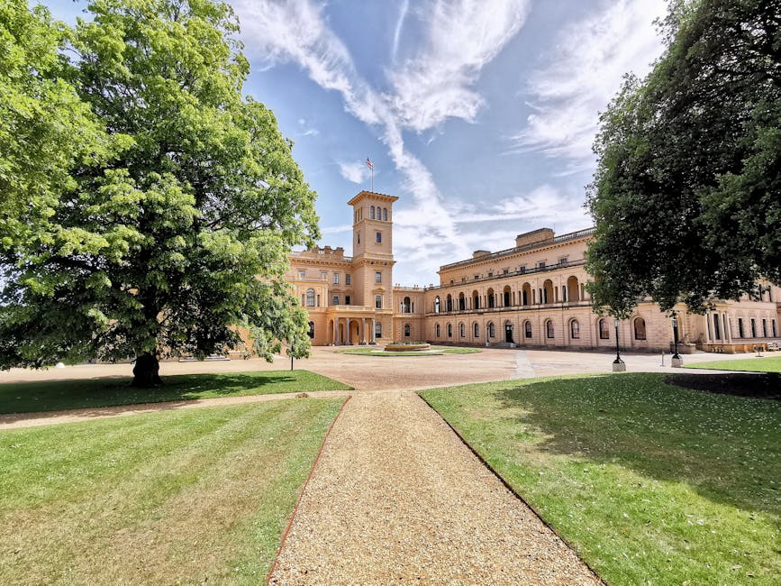 A wide view of Syon Park showing the historic palace surrounded by well-maintained green lawns and large trees, with a gravel pathway leading directly to the building's entrance. The palace's architec